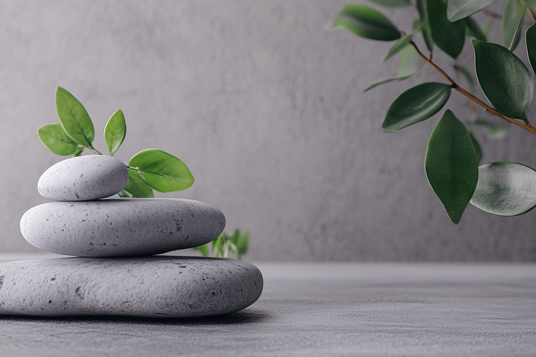 A minimalistic composition of smooth grey stones stacked in balance, surrounded by fresh green leaves. The background features a soft, neutral grey stone texture, evoking a sense of calm, wellness, and nature.